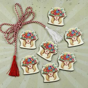 Mărțișor elegant în formă de cap de fată cocheta, cu pălărie decorată cu flori colorate, ascunzând privirea într-o aură misterioasă și rafinată.