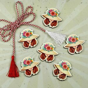 Mărțișor elegant în formă de cap de fată cocheta, cu pălărie decorată cu flori colorate, ascunzând privirea într-o aură misterioasă și rafinată.